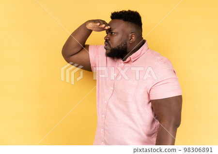 Side view of curious man with beard in pink shirt keeping palm over head and looking attentively far away, peering with expectation at long distance. Indoor studio shot isolated on yellow background. Side view of curious man with beard in pink shirt keeping palm over head and looking attentively far away, peering with expectation at long distance. Indoor studio shot isolated on yellow background. 98306891