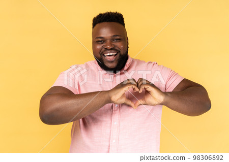 Portrait of lovely romantic happy man wearing pink shirt making heart shape with fingers, gesturing love hope charity sign, looks at camera. Indoor studio shot isolated on yellow background. Portrait of lovely romantic happy man wearing pink shirt making heart shape with fingers, gesturing love hope charity sign, looks at camera. Indoor studio shot isolated on yellow background. 98306892