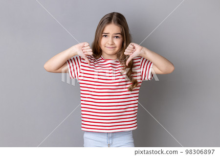 Portrait of unhappy sad little dark haired girl wearing striped T-shirt showing thumb down, expressing bad negative emotions, dislike. Indoor studio shot isolated on gray background. Portrait of unhappy sad little dark haired girl wearing striped T-shirt showing thumb down, expressing bad negative emotions, dislike. Indoor studio shot isolated on gray background. 98306897