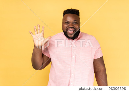 Portrait of friendly bearded man in pink shirt greeting you rising hand and waving, saying hi, glad to see you, looking at camera with toothy smile. Indoor studio shot isolated on yellow background. Portrait of friendly bearded man in pink shirt greeting you rising hand and waving, saying hi, glad to see you, looking at camera with toothy smile. Indoor studio shot isolated on yellow background. 98306898
