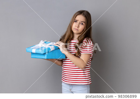 Portrait of little girl wearing striped T-shirt opening birthday gift box and looking at camera with disappointed expression, unwrapping bad present. Indoor studio shot isolated on gray background. Portrait of little girl wearing striped T-shirt opening birthday gift box and looking at camera with disappointed expression, unwrapping bad present. Indoor studio shot isolated on gray background. 98306899