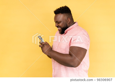 Side view of bearded happy man wearing pink shirt using mobile phone with happy expression, addicted to smartphone, texting in social network. Indoor studio shot isolated on yellow background. Side view of bearded happy man wearing pink shirt using mobile phone with happy expression, addicted to smartphone, texting in social network. Indoor studio shot isolated on yellow background. 98306907