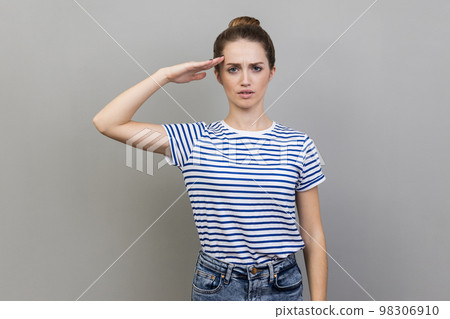 Yes, sir. Portrait of woman wearing striped T-shirt saluting with respect and responsibility, patriotic citizen listening order with honor. Indoor studio shot isolated on gray background. 98306910