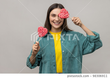 Childish woman with dark hair covering eye with heart shape lollipops, hiding, having fun with sweets, wearing casual style jacket. Indoor studio shot isolated on gray background. 98306911