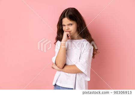 Need to think. Portrait of thoughtful little girl wearing white T-shirt holding her chin and pondering idea, confused not sure about solution. Indoor studio shot isolated on pink background. 98306913