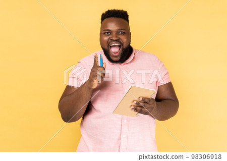 Portrait of happy man wearing pink shirt writing creative idea into notebook, drawing sketch or taking notes in paper, raised arm, keeps mouth open. Indoor studio shot isolated on yellow background. Portrait of happy man wearing pink shirt writing creative idea into notebook, drawing sketch or taking notes in paper, raised arm, keeps mouth open. Indoor studio shot isolated on yellow background. 98306918
