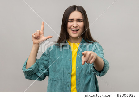 Disrespectful rude young woman showing loser gesture and pointing on you, abuser, mocking your failures, wearing casual style jacket. Indoor studio shot isolated on gray background. 98306923