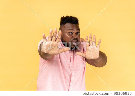 No, it's scary. Portrait of frightened shocked man wearing pink shirt raising hands in fear, looking horrified and panicking, hiding from phobia. Indoor studio shot isolated on yellow background. No, it's scary. Portrait of frightened shocked man wearing pink shirt raising hands in fear, looking horrified and panicking, hiding from phobia. Indoor studio shot isolated on yellow background. 98306953