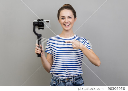 Portrait of happy smiling stylish woman wearing striped T-shirt holding mobile phone, pointing at steadicam, looking at camera with positive expression. Indoor studio shot isolated on gray background. 98306958