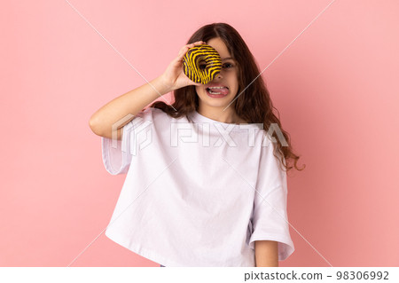 Portrait of joyful adorable little girl wearing white T-shirt standing having fun, holding and covering eye with donut and showing tongue out. Indoor studio shot isolated on pink background. Portrait of joyful adorable little girl wearing white T-shirt standing having fun, holding and covering eye with donut and showing tongue out. Indoor studio shot isolated on pink background. 98306992