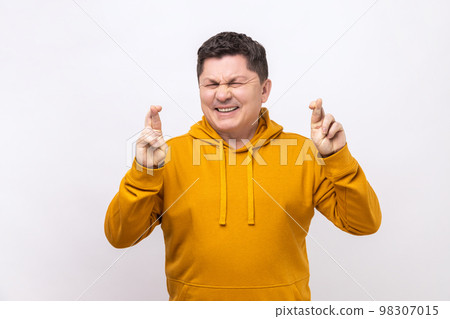 Portrait of middle aged man crossing fingers and closing eyes making wish, believing in miracle, hope in success, wearing urban style hoodie. Indoor studio shot isolated on white background. 98307015