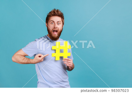 Portrait of surprised handsome bearded man pointing finger on hashtag in his hand with open mouth and big eyes, website search. Indoor studio shot isolated on blue background. 98307023