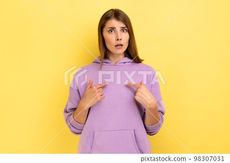 Portrait of amazed woman pointing herself and looking at camera with shocked expression, astonished by sudden success, wearing purple hoodie. Indoor studio shot isolated on yellow background. 98307031