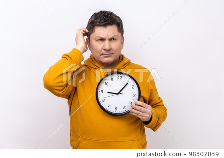 Portrait of middle aged man rubbing his head holding in hands big wallclock, has no time, worried about deadline, wearing urban style hoodie. Indoor studio shot isolated on white background. 98307039