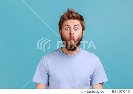 Portrait of funny comic face handsome bearded man standing with fish lips and looking at camera, childish behavior, having fun, good mood. Indoor studio shot isolated on blue background. Portrait of funny comic face handsome bearded man standing with fish lips and looking at camera, childish behavior, having fun, good mood. Indoor studio shot isolated on blue background. 98307040