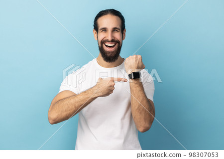 Look at clock, no rush. Portrait of man with beard wearing white T-shirt pointing wrist watch and expressing optimism about time, not busy. Indoor studio shot isolated on blue background. 98307053