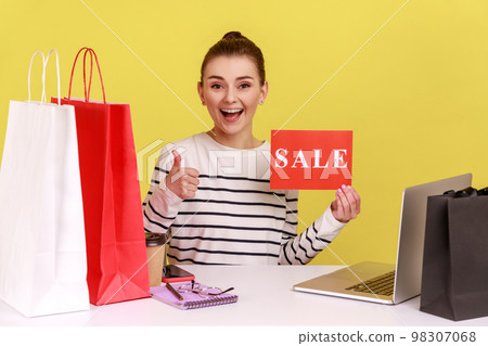 Excited woman office worker showing thumb up holding card with sale inscription, satisfied with shopping, sitting at workplace with paper bags. Indoor studio studio shot isolated on yellow background. 98307068