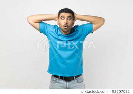 Portrait of shocked surprised man wearing blue T- shirt standing looking at camera with big eyes, keeps hands behind head, sees something astonished. Indoor studio shot isolated on gray background. 98307081