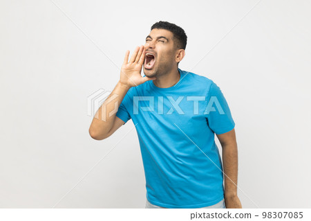 Portrait of excited handsome attractive unshaven man wearing blue T- shirt standing keeps hands near opened mouth, screaming news. Indoor studio shot isolated on gray background. 98307085