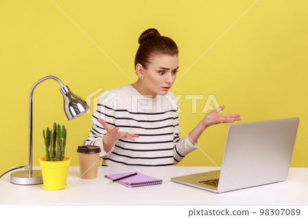 Young woman office worker raising hands in indignant gesture, asking why and looking angrily while having video call with business partner. Indoor studio studio shot isolated on yellow background. Young woman office worker raising hands in indignant gesture, asking why and looking angrily while having video call with business partner. Indoor studio studio shot isolated on yellow background. 98307089