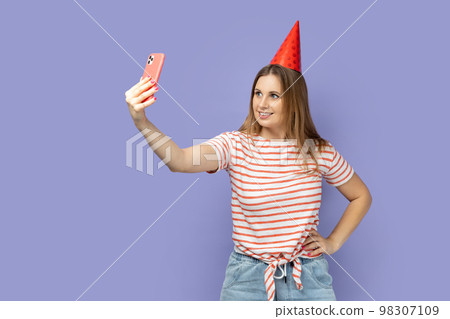 Portrait of blond woman blogger wearing striped T-shirt and party cone broadcasting livestream or taking selfie for her blog, keeps hand on hip. Indoor studio shot isolated on purple background. Portrait of blond woman blogger wearing striped T-shirt and party cone broadcasting livestream or taking selfie for her blog, keeps hand on hip. Indoor studio shot isolated on purple background. 98307109