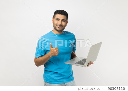 Portrait of happy delighted attractive unshaven man wearing blue T- shirt standing holding laptop in hands, working online, showing thumb up. Indoor studio shot isolated on gray background. Portrait of happy delighted attractive unshaven man wearing blue T- shirt standing holding laptop in hands, working online, showing thumb up. Indoor studio shot isolated on gray background. 98307110