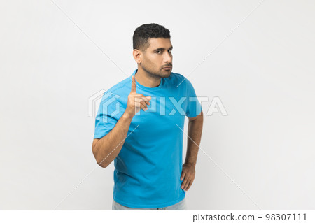 Portrait of serious bossy attractive unshaven man wearing blue T- shirt standing with raised index finger, warning somebody, showing disapproval gesture. Indoor studio shot isolated on gray background Portrait of serious bossy attractive unshaven man wearing blue T- shirt standing with raised index finger, warning somebody, showing disapproval gesture. Indoor studio shot isolated on gray background 98307111