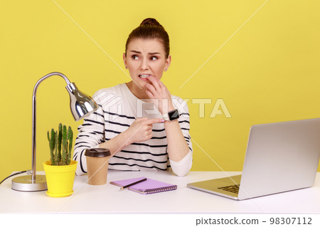 Too late, no time to meet deadline. Worried woman secretary sitting at workplace, looking at wrist watch with anxiety, nervous about late hour. Indoor studio studio shot isolated on yellow background. Too late, no time to meet deadline. Worried woman secretary sitting at workplace, looking at wrist watch with anxiety, nervous about late hour. Indoor studio studio shot isolated on yellow background. 98307112