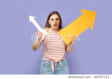 Portrait of shocked scared blond woman wearing striped T-shirt holding two arrows indicating to different sizes, looking at camera with big eyes. Indoor studio shot isolated on purple background. Portrait of shocked scared blond woman wearing striped T-shirt holding two arrows indicating to different sizes, looking at camera with big eyes. Indoor studio shot isolated on purple background. 98307123