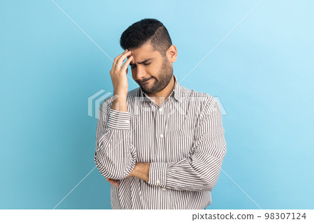 Businessman with beard making facepalm gesture keeping hand on head, blaming himself for bad memory, unforgivable mistake, wearing striped shirt. Indoor studio shot isolated on blue background. Businessman with beard making facepalm gesture keeping hand on head, blaming himself for bad memory, unforgivable mistake, wearing striped shirt. Indoor studio shot isolated on blue background. 98307124