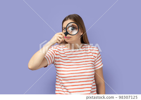 Portrait of funny positive blond woman wearing striped T-shirt standing, holding magnifying glass and looking at camera with big zoom eye. Indoor studio shot isolated on purple background. Portrait of funny positive blond woman wearing striped T-shirt standing, holding magnifying glass and looking at camera with big zoom eye. Indoor studio shot isolated on purple background. 98307125