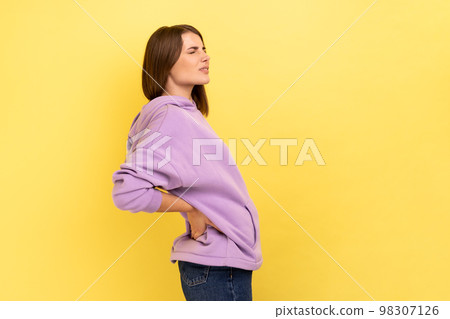 Profile portrait of unhealthy frustrated anxious woman holding hands on lower back, kidney injury, backache, wearing purple hoodie. Indoor studio shot isolated on yellow background. 98307126