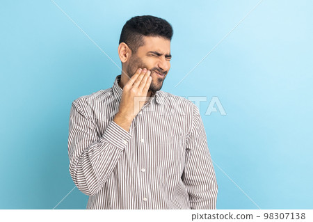 Unhappy bearded businessman feeling toothache, touching sore cheek, suffering from cavities, cracked teeth, gum recession, wearing striped shirt. Indoor studio shot isolated on blue background. 98307138