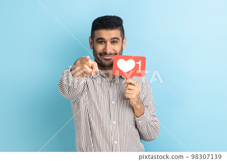 Do you rate my post in social network. Smiling man blogger holding like counter sign and pointing finger at camera, wearing striped shirt. Indoor studio shot isolated on blue background. 98307139