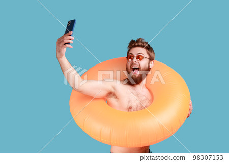 Portrait of excited bearded man posing with orange rubber ring and holding mobile phone, making selfie, enjoying his vacation at seaside. Indoor studio shot isolated on blue background. 98307153