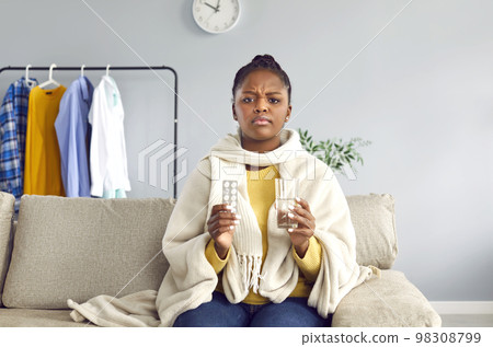 Sick, sad young woman sitting on sofa at home, holding glass of water and medicine 98308799