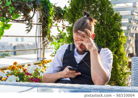 Upset man looking at smartphone holding his head while sitting in penthouse got bad news Upset man looking at smartphone holding his head while sitting in penthouse got bad news 98310004