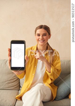 Woman Showing Smartphone At Home. Girl Sitting On Sofa And Demonstrating Mobile Woman Showing Smartphone At Home. Girl Sitting On Sofa And Demonstrating Mobile 98311517