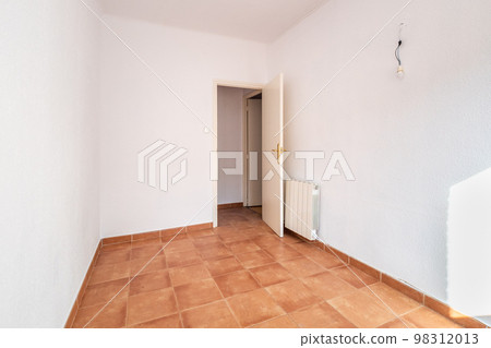 An empty spacious room with an open door, white walls and ceiling, caramel-colored square floor tiles and a radiator on the wall for warmth in the cold season. The room is lit by bright daylight. 98312013