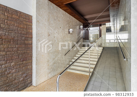Entrance of a house with long corridor, stairs to elevator area with ramp for disabled person wheelchair, brick and tiled walls, metal mailboxes. Entrance of a house with long corridor, stairs to elevator area with ramp for disabled person wheelchair, brick and tiled walls, metal mailboxes. 98312933