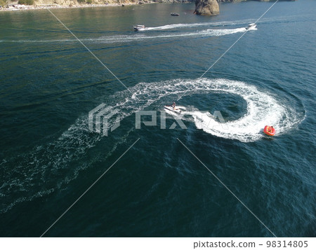 Happy people swim on air mattress behind a high speed water bike. Tourists ride on inflatable watercraft boat. Sea attraction. Water scooter skating people on large Inflatable raft. Aerial copter view Happy people swim on air mattress behind a high speed water bike. Tourists ride on inflatable watercraft boat. Sea attraction. Water scooter skating people on large Inflatable raft. Aerial copter view 98314805