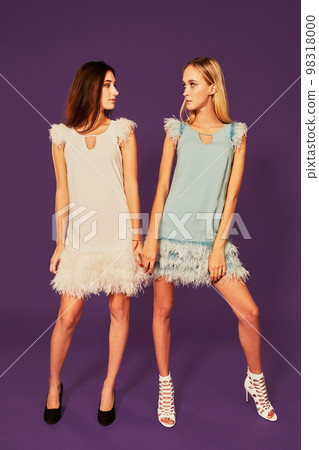 Professional photo of two young women, blonde and brunette, in cocktail dresses posing on purlpe background Professional photo of two young women, blonde and brunette, in cocktail dresses posing on purlpe background 98318000