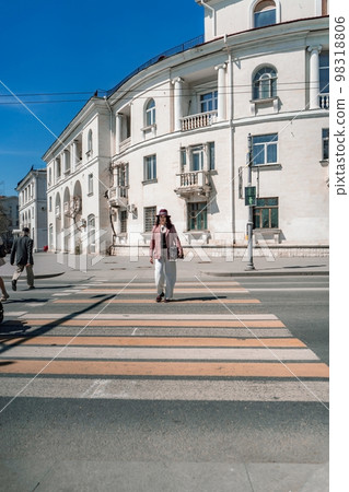 Woman city road crossing. Stylish woman in a hat crosses the road at a pedestrian crossing in the city. Dressed in white trousers and a jacket with a bag in her hands. 98318806