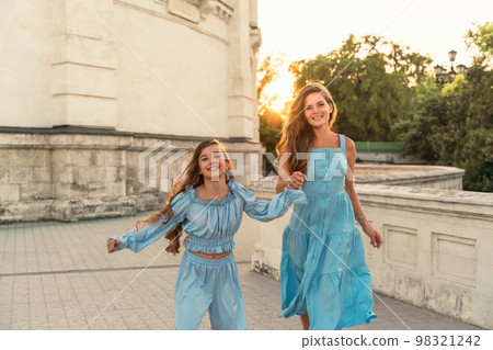 Portrait of a mother and daughter in blue dresses with flowing long hair against the backdrop of a sunset and a white building. They run and smile at the camera. Family stories on the weekend 98321242