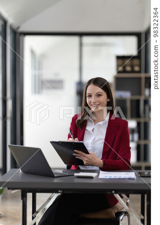 Portrait smiling woman sitting at desk working on laptop taking notes in notebook, happy millennial female working online, watching webinar using computer and writing check list 98322364