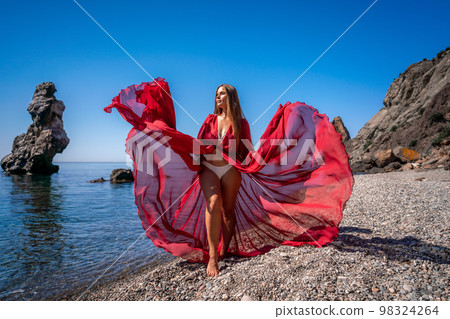 Beautiful sensual woman in a flying red dress and with long hair, stands on the seashore Beautiful sensual woman in a flying red dress and with long hair, stands on the seashore 98324264