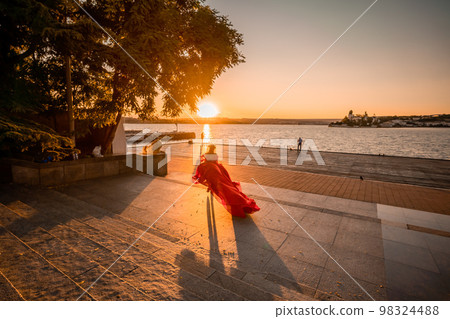 Sunrise red dress. A woman in a long red dress against the backdrop of sunrise, bright golden light of the sun's rays. The concept of femininity, harmony. 98324488