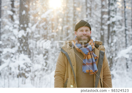 Portrait of young bearded man in warm clothing smiling at camera standing in winter forest Portrait of young bearded man in warm clothing smiling at camera standing in winter forest 98324523