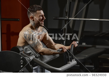Portrait of young muscular man training shirtless in gym indoors. Hand exercises. Workout. Concept of sport, workout, strength Portrait of young muscular man training shirtless in gym indoors. Hand exercises. Workout. Concept of sport, workout, strength 98324930