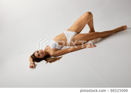 Femininity. Slim body. Portrait of young beautiful girl posing in white underwear over grey studio background. Concept of body and skin care, fitness 98325006